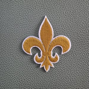 Puede incluir: Parche bordado de flor de lis dorada con borde blanco. El parche presenta un diseño detallado de la flor de lis estilizada, un símbolo de la realeza. El parche es probablemente para fines decorativos, como en ropa o accesorios.
