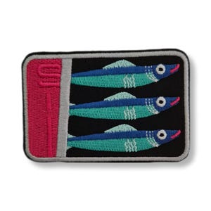 Peut inclure: Écusson brodé représentant trois poissons dans une boîte de sardines. Les poissons sont bleus et turquoise avec des bouches roses et des détails blancs, sur fond noir. La boîte est rose avec un bord argenté.