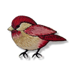 Puede incluir: Parche bordado de un pájaro con cabeza y alas rojas, cuerpo beige y patas negras. El pájaro mira hacia la izquierda, con el pico ligeramente abierto. El parche está sobre un fondo blanco.