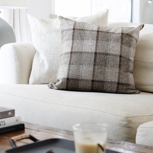 May include: A white couch with two pillows, one with a plaid pattern and one with a textured pattern. A wooden coffee table with a black tray, a glass of iced coffee, and a stack of books with the titles "down to earth", "wonderplants", and "William Garnett".