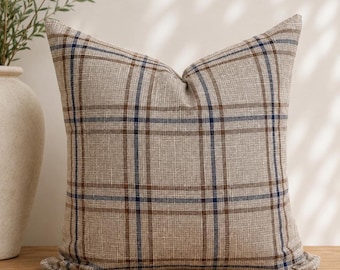 Peut inclure: Un coussin carré doté d'une base en tissu beige tissé avec un motif à carreaux prononcé. Le design se compose de lignes horizontales et verticales entrecroisées dans des tons de brun foncé et de bleu marine, créant une grille structurée.