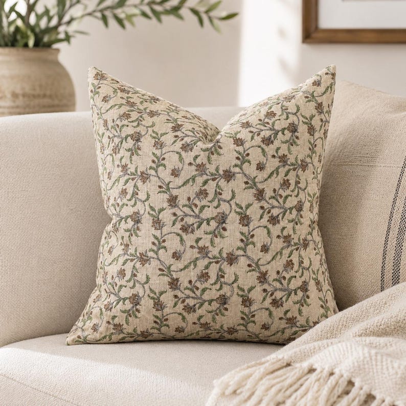 May include: Square throw pillow with a floral pattern. The pillow features a beige background with a repeating design of brown and green flowers and vines. The pillow is placed on a beige sofa.