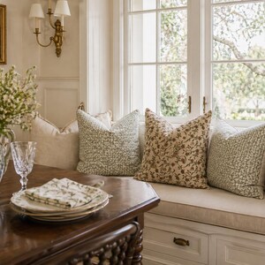 May include: A cozy interior scene featuring a dark wood table set with plates, napkins, and a water glass. A blue and white floral pitcher holds flowers. Pillows with floral and textured patterns adorn a window seat.