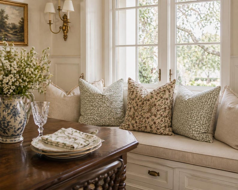 May include: A cozy interior scene featuring a dark wood table set with plates, napkins, and a water glass. A blue and white floral pitcher holds flowers. Pillows with floral and textured patterns adorn a window seat.