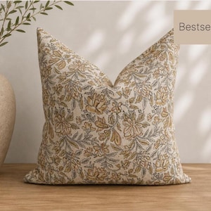 May include: A square throw pillow featuring a cream-colored fabric with a repeating floral pattern in muted mustard yellow and charcoal gray. The pillow has a label in the top right corner with the text Bestseller printed in black.