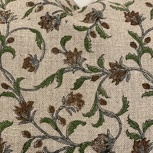 May include: Close-up of a beige pillow with a floral pattern. The design features brown flowers, green leaves, and gray stems. The fabric has a textured appearance, suggesting a linen or similar material.