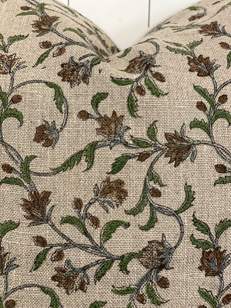 May include: Close-up of a beige pillow with a floral pattern. The design features brown flowers, green leaves, and gray stems. The fabric has a textured appearance, suggesting a linen or similar material.