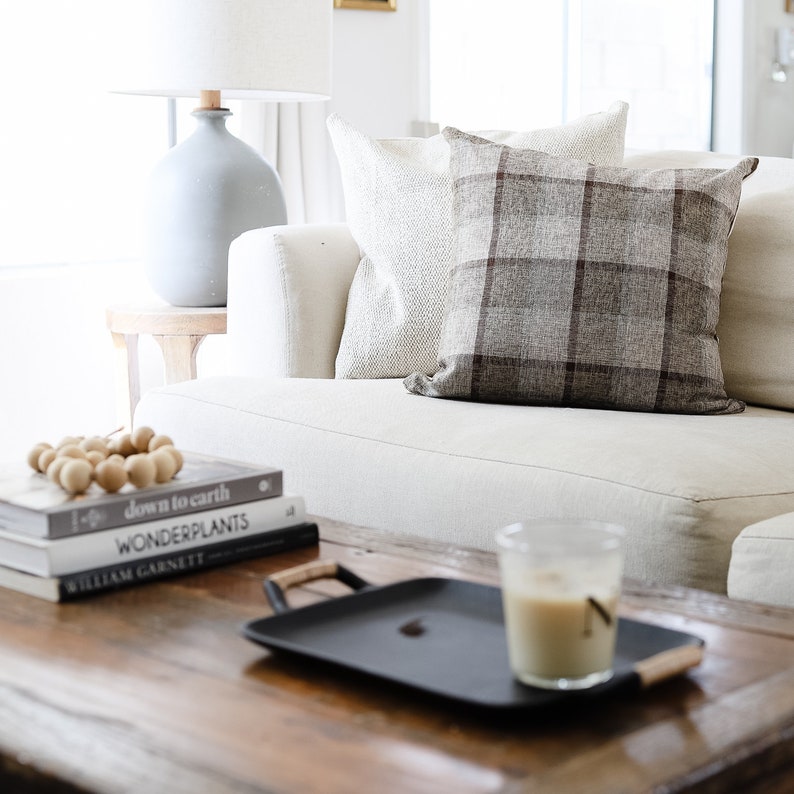 May include: A white couch with two pillows, one with a plaid pattern and one with a textured pattern. A wooden coffee table with a black tray, a glass of iced coffee, and a stack of books with the titles "down to earth", "wonderplants", and "William Garnett".