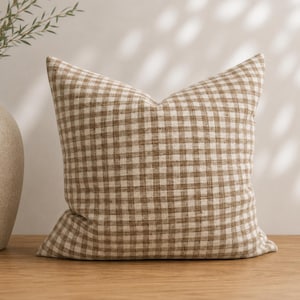 May include: A square throw pillow featuring a brown and white gingham check pattern. The fabric has a visible woven texture, and the pillow is plumped to a firm shape. The design consists of a repeating grid of small, alternating brown and white squares.