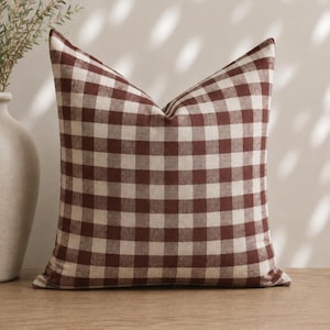 May include: A square throw pillow featuring a classic gingham pattern in deep brown and off-white. The fabric displays a visible woven texture throughout the checkered design, creating a consistent grid of alternating dark and light squares.