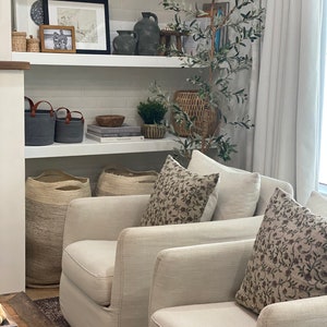 May include: A living room scene with two cream-colored armchairs, adorned with floral patterned pillows. A bookshelf displays decorative items, including vases, baskets, and framed artwork. A faux olive tree adds a touch of nature to the space.