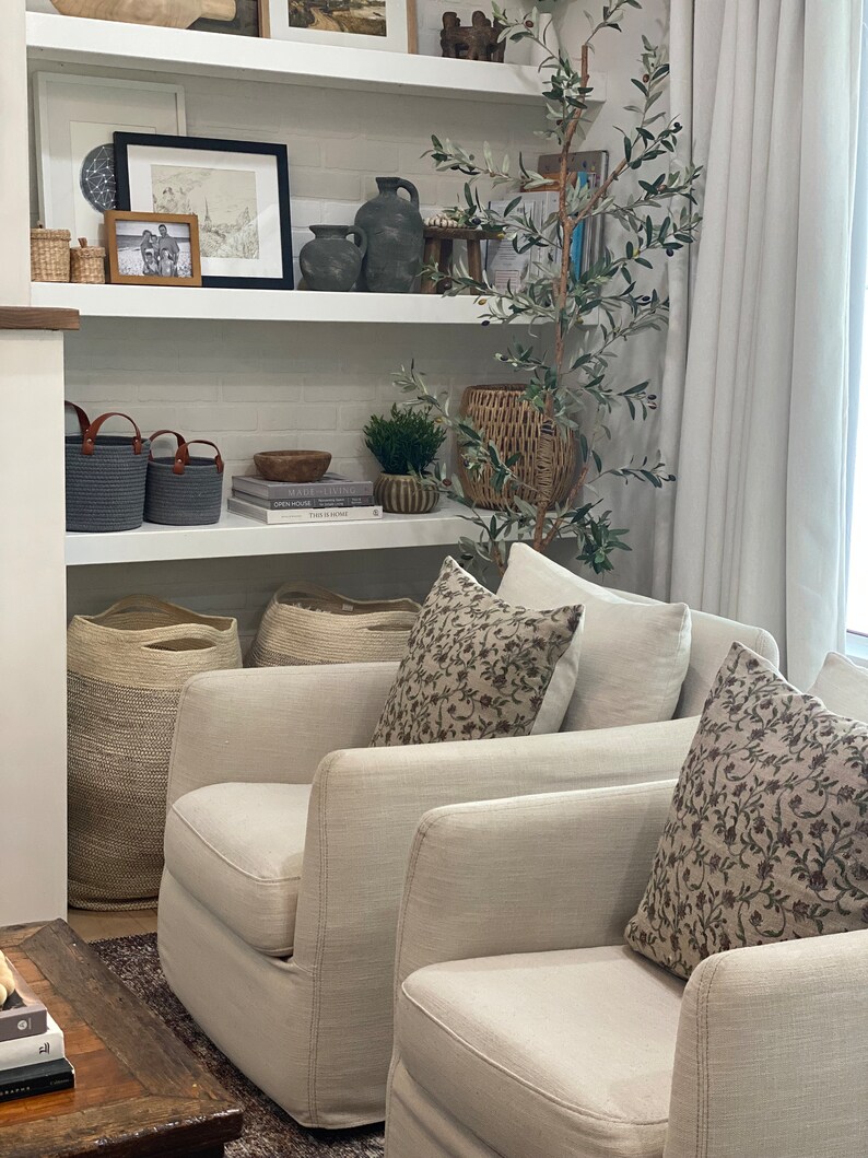 May include: A living room scene with two cream-colored armchairs, adorned with floral patterned pillows. A bookshelf displays decorative items, including vases, baskets, and framed artwork. A faux olive tree adds a touch of nature to the space.