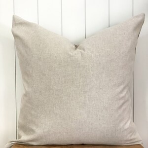 May include: A large, square, beige pillow with a textured fabric. The pillow is resting on a rustic wooden stool against a white-painted wood-paneled wall. The pillow has a simple, minimalist design.