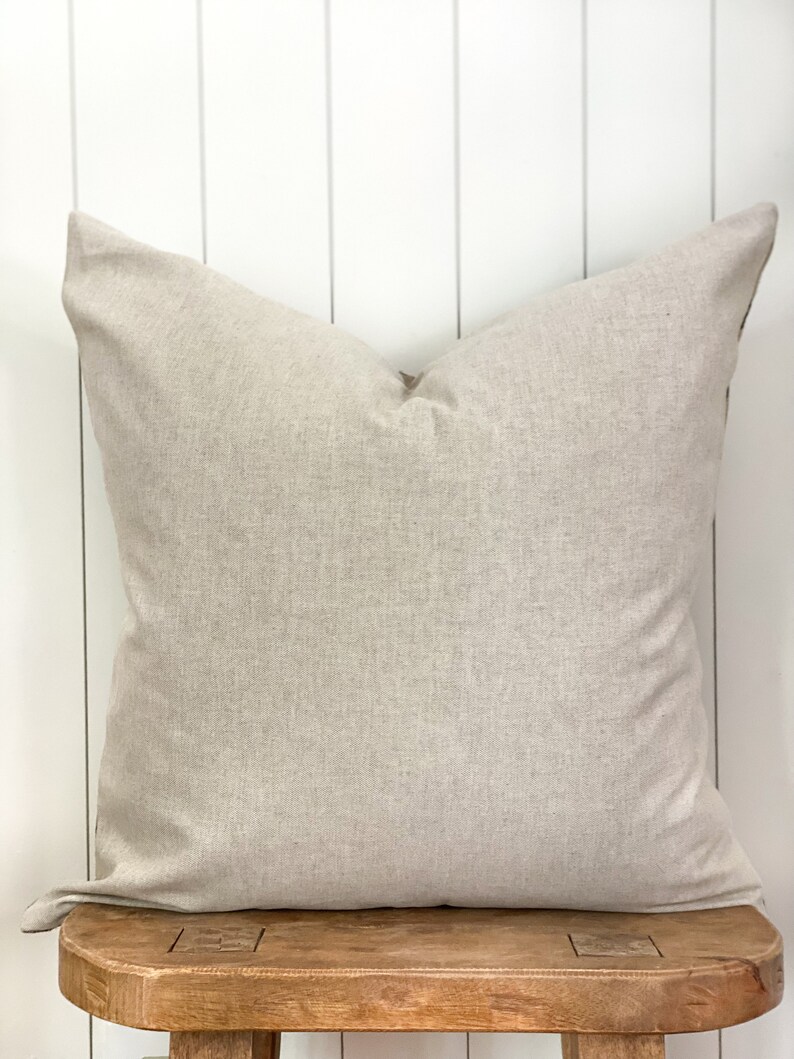 May include: A large, square, beige pillow with a textured fabric. The pillow is resting on a rustic wooden stool against a white-painted wood-paneled wall. The pillow has a simple, minimalist design.