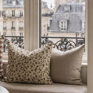 May include: Two decorative throw pillows on a window seat. One pillow features a floral pattern in brown and green on a cream background. The other pillow is a solid beige color. The window overlooks a city.
