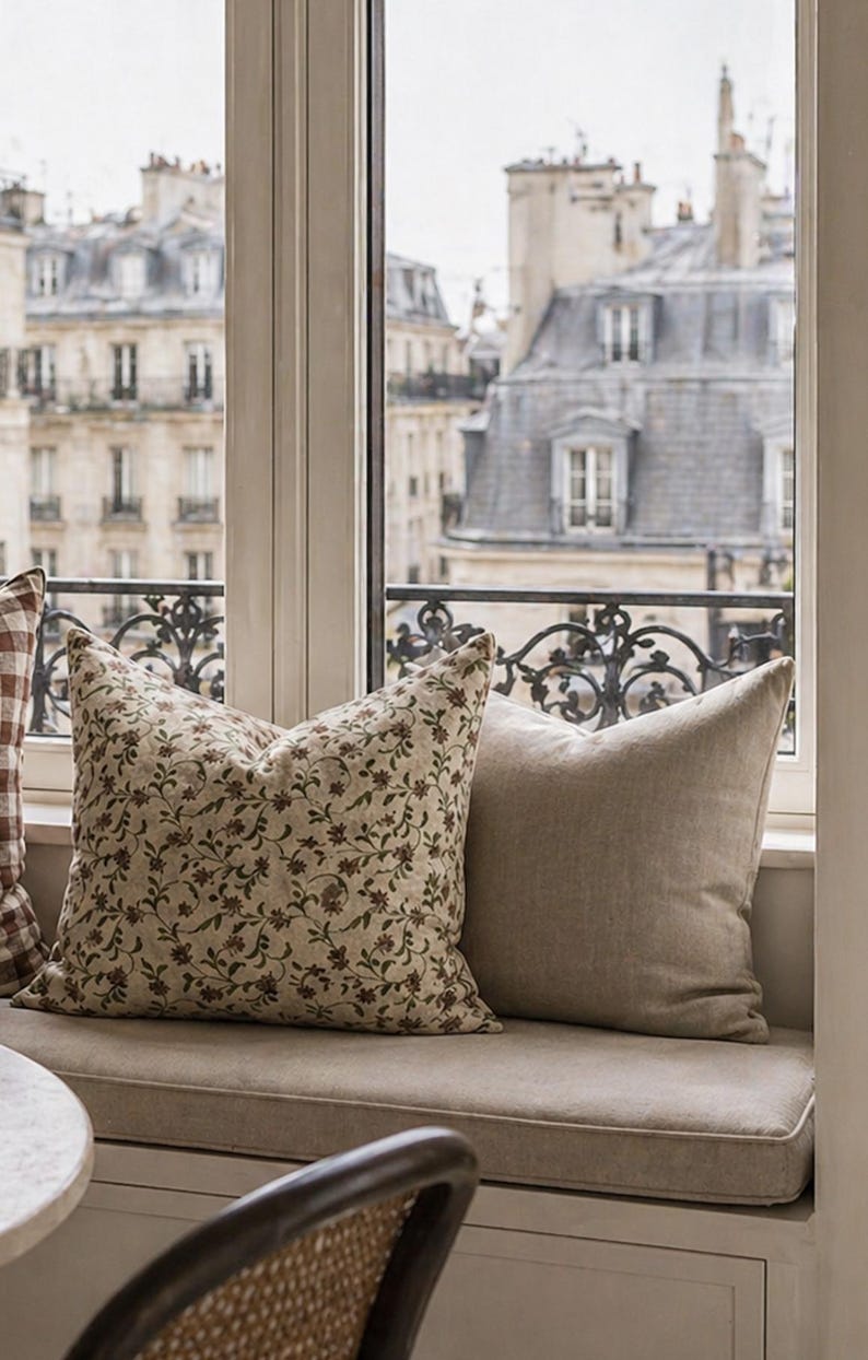 May include: Two decorative throw pillows on a window seat. One pillow features a floral pattern in brown and green on a cream background. The other pillow is a solid beige color. The window overlooks a city.