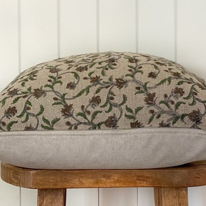 May include: A rectangular pillow with a floral pattern. The top of the pillow features a beige fabric with a repeating pattern of brown flowers and green leaves. The bottom is a solid light gray. The pillow is resting on a small wooden stool.
