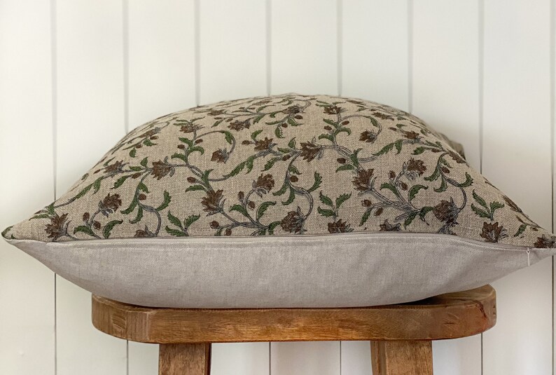 May include: A rectangular pillow with a floral pattern. The top of the pillow features a beige fabric with a repeating pattern of brown flowers and green leaves. The bottom is a solid light gray. The pillow is resting on a small wooden stool.