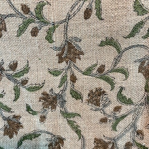 May include: Close-up of a beige fabric with a repeating floral pattern. The design features brown flowers, green leaves, and gray stems. The fabric has a textured, woven appearance, suggesting a natural fiber like linen or burlap.
