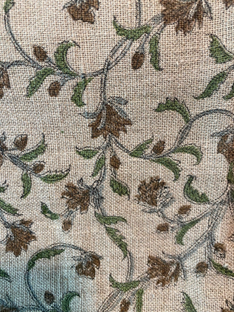 May include: Close-up of a beige fabric with a repeating floral pattern. The design features brown flowers, green leaves, and gray stems. The fabric has a textured, woven appearance, suggesting a natural fiber like linen or burlap.