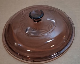 May include: A round, brown glass lid with a black knob. The lid has a slightly domed shape with concentric ridges radiating from the center. The glass is transparent, allowing a view of the contents below.