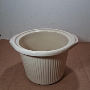 May include: A cream-colored ceramic slow cooker insert with a round opening and a wide, flat rim. The exterior features a vertical ribbed texture, and the interior is smooth. Two small, integrated handles extend from opposite sides of the rim.