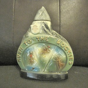 May include: A green ceramic decanter shaped like a mountain with a white peak. The decanter has a gold inscription that reads "Rush to the Rockies". The decanter features three scenes: a skier, a person on a sled, and a deer.