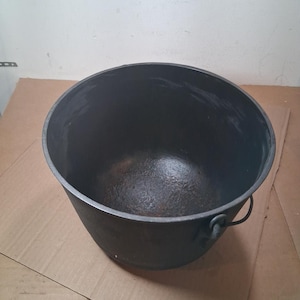 May include: A large, round, black cast iron cauldron with a wide open top and a curved metal handle attached to the sides. The interior surface shows a dark, textured finish with signs of wear, while the exterior is a solid, matte black color.