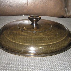 May include: A brown glass lid with a knob on top. The lid is round and has a slightly domed shape.