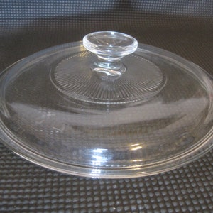 May include: Clear glass lid with a knob on top. The lid has a raised, circular design around the edge.
