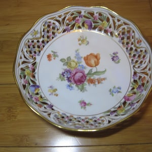 Puede incluir: Un plato de porcelana blanca con un borde dorado y un diseño de corte intrincado. El plato presenta un motivo floral pintado a mano con flores rosas, amarillas, naranjas y azules.