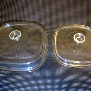 May include: Two clear glass lids with rounded square shapes and central circular knobs. The lids feature a raised rim around the perimeter and a smooth, transparent surface, designed to fit cookware of two different sizes.
