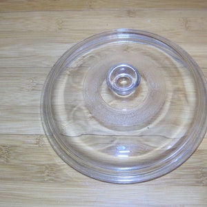 May include: A clear glass lid with a knob on top. The lid is round and has a slightly raised edge.