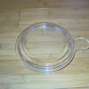 Könnte beinhalten: Ein klarer Glasdeckel mit Griff. Der Deckel ist kreisförmig mit einem leicht erhöhten Rand und einem kleinen, gebogenen Griff an einer Seite. Der Deckel ist für einen runden Behälter konzipiert.