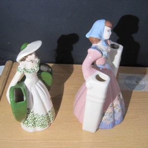 Puede incluir: Dos figuritas de cerámica vintage de mujeres con vestidos. Una lleva un sombrero verde y sostiene un jarrón verde. La otra lleva un sombrero azul y sostiene un jarrón rosa.