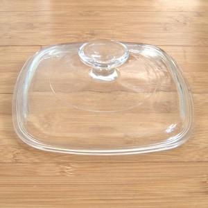 Op de afbeelding: Een helder, vierkant glazen deksel met een afgeronde handgreep. Het deksel is ontworpen voor kookgerei en ligt op een lichtbruin houten oppervlak. Het transparante ontwerp maakt de inhoud zichtbaar.