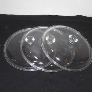 May include: Three clear glass pot lids with small, round handles. The lids are circular with a slightly raised rim and are arranged on a black surface. The handles are clear and appear to be made of glass.
