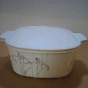 May include: An off-white, oval-shaped casserole dish with a matching lid. The dish features a floral design with pink and green accents. The lid has two handles for easy handling. A practical kitchen item.