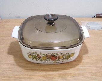 May include: White ceramic casserole dish with a brown glass lid. The dish has a floral design with vegetables and the text "La  Marjolaine".