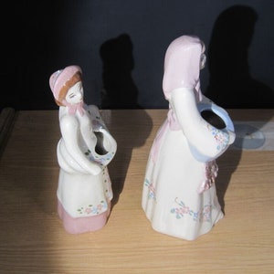 Puede incluir: Dos figuritas de cerámica de mujeres con vestidos con motivos florales. Ambas figuritas sostienen cestas con diseños florales. Las figuritas están pintadas en tonos de rosa, azul y blanco.