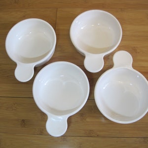 May include: Four white ceramic bowls with a handle on a light brown wooden surface. The bowls are round with a small, rounded handle extending from one side. The bowls are empty and appear to be new.