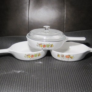 May include: A set of three white ceramic cookware pieces with a colorful fruit and vegetable design. One piece has a clear glass lid. The text "Le Basil" is printed on the front of each piece.