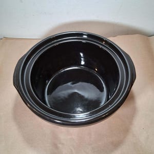 May include: A round, black ceramic slow cooker insert with a glossy finish. The vessel features a wide, open top with a thick, stepped rim and two integrated side handles. The interior is smooth, deep, and dark, reflecting light on its surface.