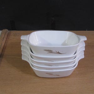 Puede incluir: Una pila de cinco fuentes blancas para hornear, de forma cuadrada, con asas. Cada fuente presenta un diseño de espiga de trigo marrón. Las fuentes están apiladas sobre una superficie de madera marrón claro.