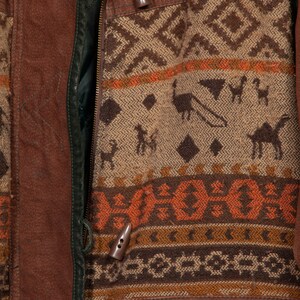 May include: Close-up of a brown leather jacket with a zipper closure and a patterned fabric panel. The fabric panel features a geometric design in shades of brown, orange, and white. The jacket has two decorative buttons.