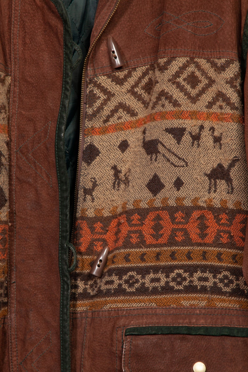 May include: Close-up of a brown leather jacket with a zipper closure and a patterned fabric panel. The fabric panel features a geometric design in shades of brown, orange, and white. The jacket has two decorative buttons.