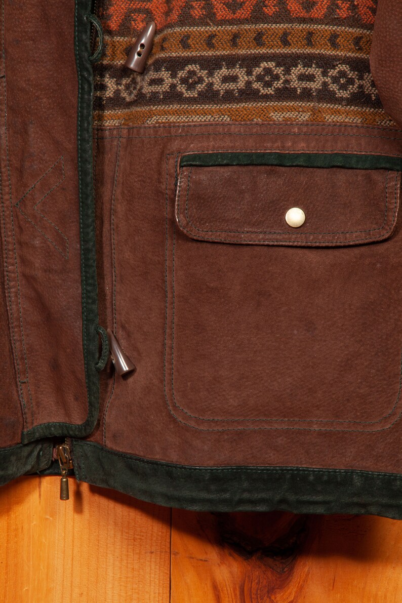 May include: A brown leather jacket with a green suede trim and a zipper closure. The jacket has a large pocket on the front and a patterned fabric panel on the top.