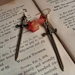 May include: A pair of earrings featuring a miniature sword charm and a red, translucent dice charm. The earrings are on a book with text about discounting.