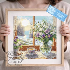 May include: A framed cross-stitch artwork depicting a winter scene with a window view. The scene includes a vase of flowers, a teacup, and a snowy landscape. The artwork is in a light wood frame and features the text "Your Family Printo Cross Stitch".