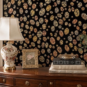 May include: A black wallpaper featuring a repeating pattern of metallic gold and bronze oval shapes. The wall is decorated with a floral lamp, a gold picture frame, a stack of books labeled Tilstoy Anna Karenina, and a vase of flowers.